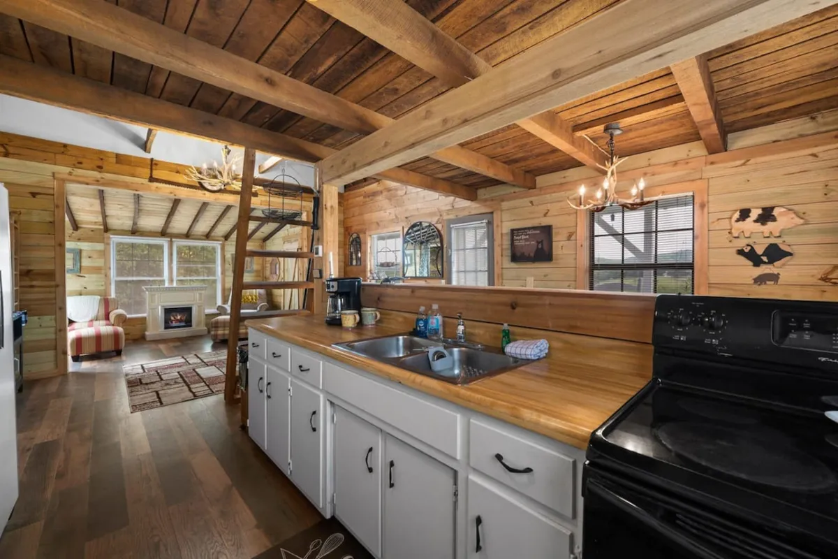 The image centers on the kitchen counter and sink, with wooden beams overhead and the stove nearby. The living area with a fireplace is visible in the background, hinting at seamless flow between spaces. The atmosphere is warm and social, highlighting people gathering around the kitchen island and sharing meals.