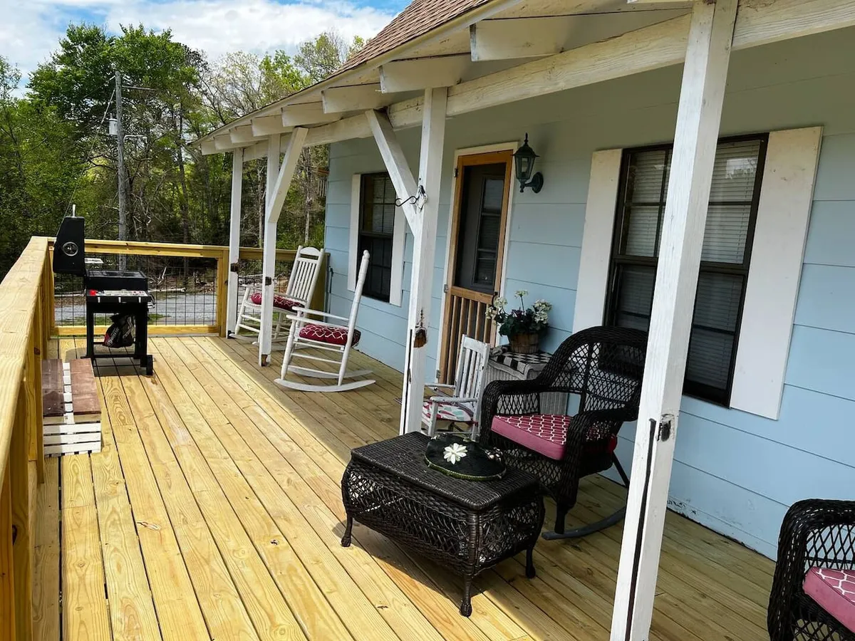 A cozy porch setting featuring comfortable rocking chairs, a grill, a coffee table adorned with flowers, and decorative plants. The porch looks out onto a serene, leafy setting—ideal for morning coffee or evening gatherings, with touches of Southern hospitality.