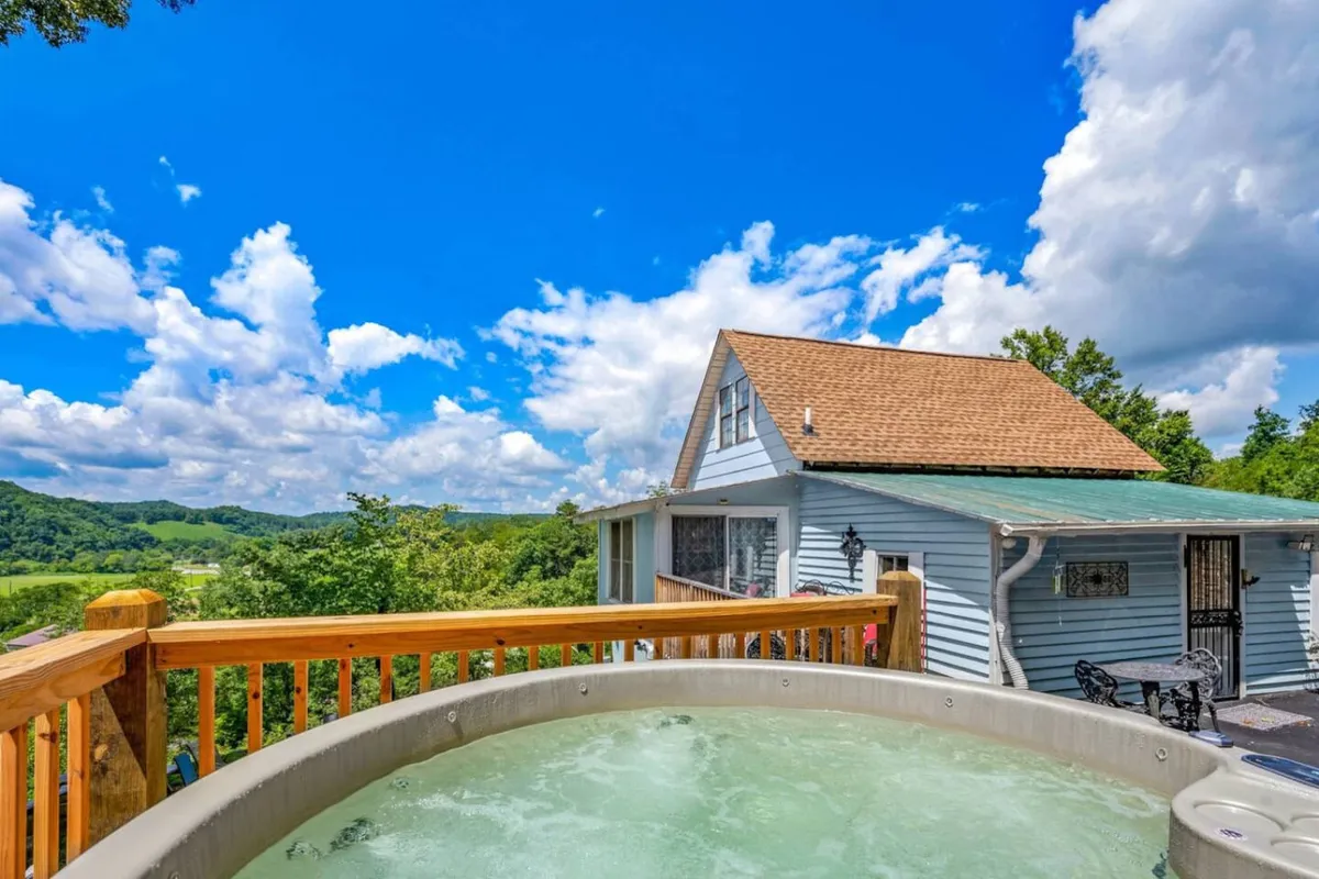 A hot tub sits on a deck overlooking a scenic mountain view with blue skies and clouds above. The inviting setup suggests relaxing soaks in all seasons, whether after a winter day in the snow or a summer evening admiring sunsets and stargazing.