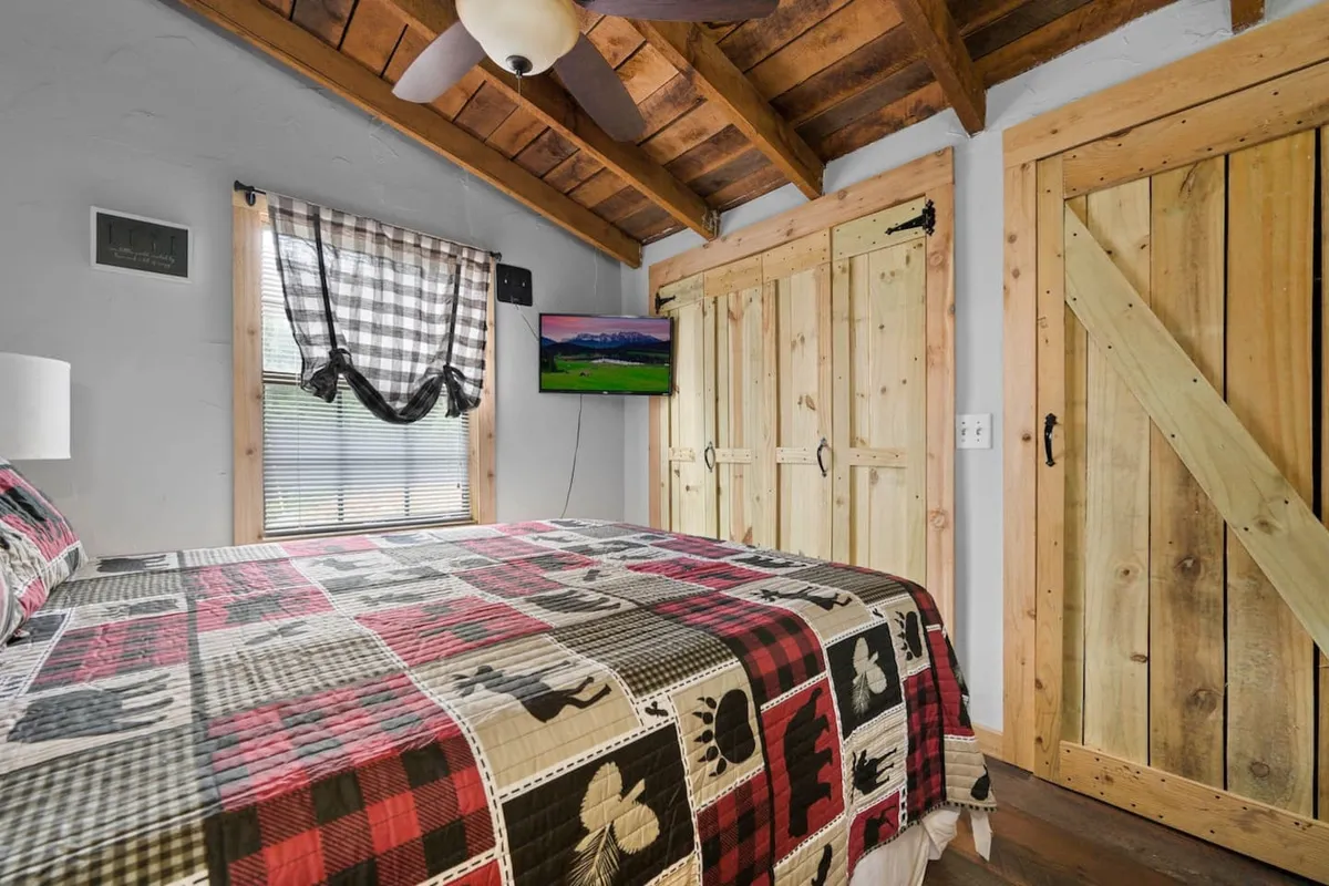 A warmly lit bedroom features a comfortable bed with a patchwork quilt, rustic wooden sliding doors, a ceiling fan with light, and a wall-mounted TV. The inviting space exudes cabin charm and offers a cozy, restful setting.