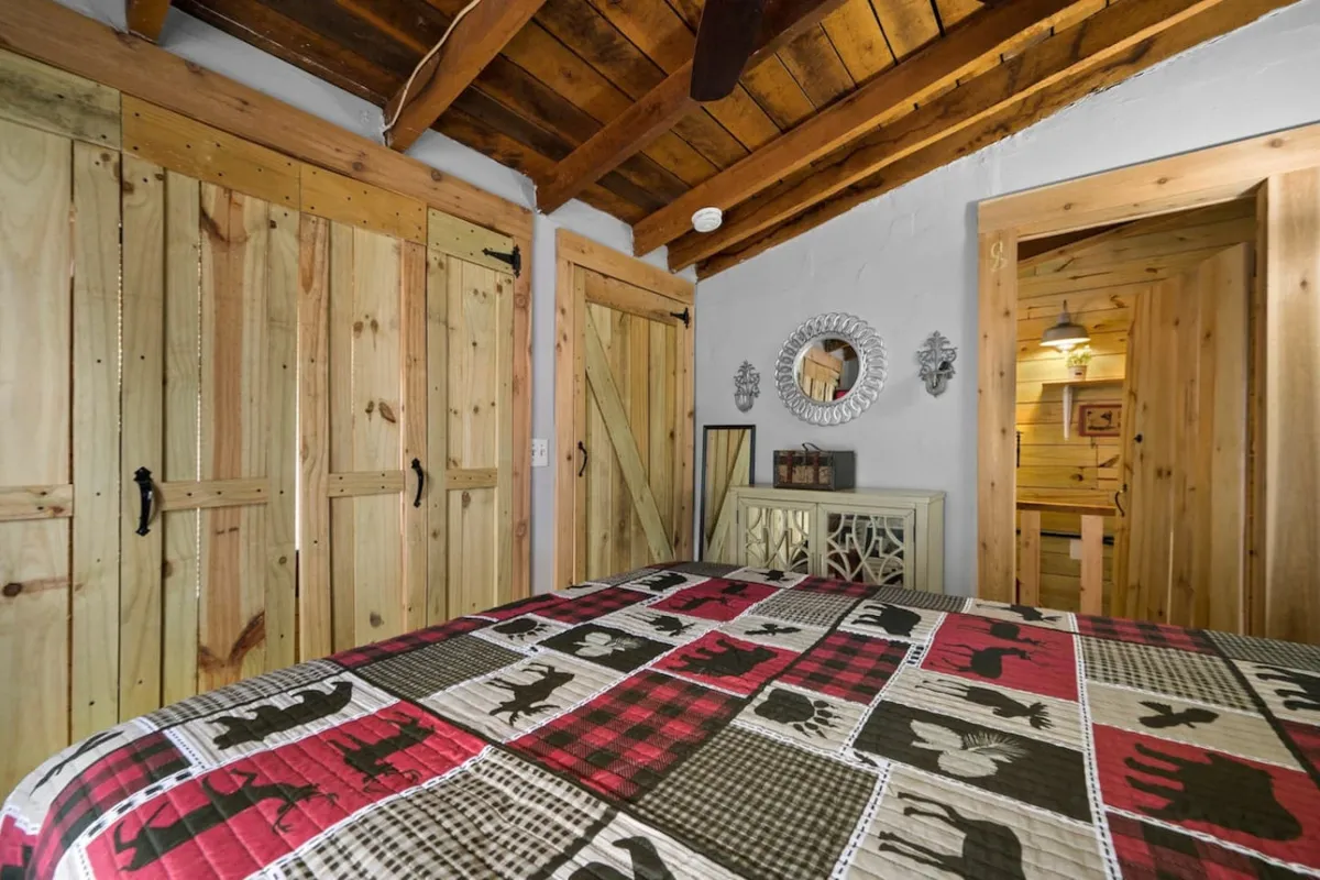 A spacious bedroom showcases a patterned quilt on the bed, rustic wooden barn doors, and a round mirror on the wall. The room feels intimate yet large enough for families, with plenty of natural light.
