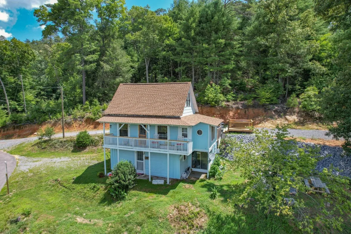 This scene features the blue house nestled among trees, highlighting the balcony, driveway, and surrounding natural landscape. The image suggests easy access to nearby trails and the welcoming outdoor environment.