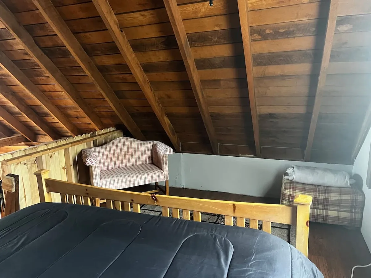 A rustic bedroom scene with a wooden bed frame, plaid armchair, and angled wooden beams. The window lets in natural light, creating a bright and tranquil atmosphere ideal for morning relaxation or late-night reading.