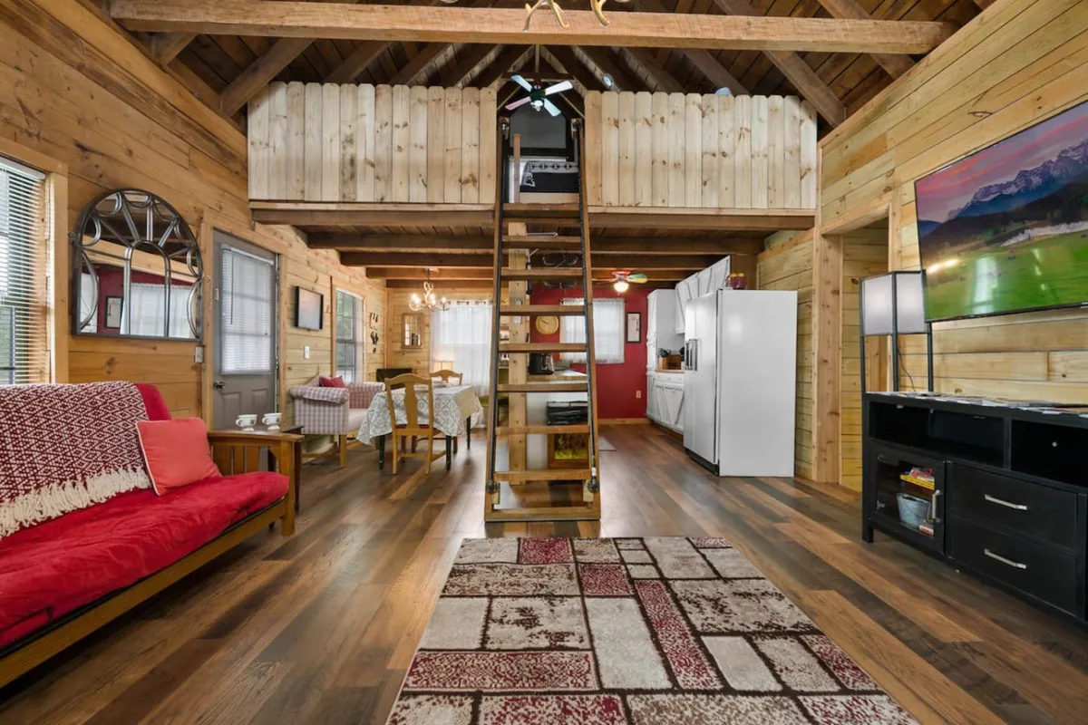 This photo captures a wide view of the open-plan living room, kitchen, and dining space. The focal points include a plush sofa arranged around an electric fireplace, a large wooden dining table ready for group meals, kitchen appliances for easy meal prep, and a prominent TV mounted for entertainment. Warm lighting and natural wood accents create a cozy, inviting ambiance.