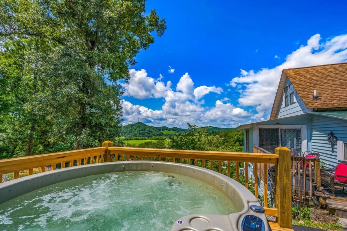 A serene outdoor hot tub set on a deck with distant mountain views, surrounded by green trees and brilliant blue sky. The scene radiates tranquility, offering a secluded spot for couples to unwind, soak, and gaze at the sunset or stars.