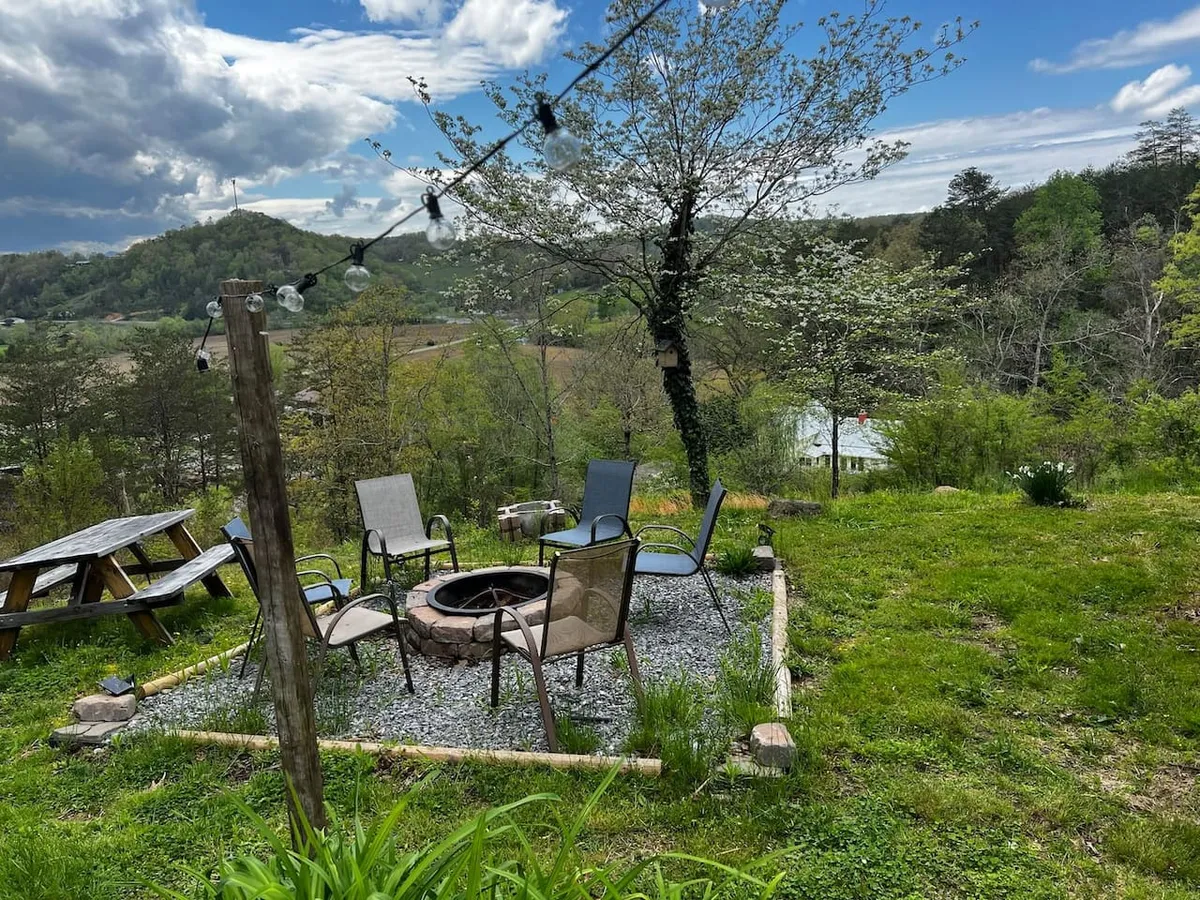 A cozy fire pit surrounded by comfortable chairs and a picnic table, all set against rolling hills and hanging string lights. The soft lighting and distant hills evoke the magic of gathering at dusk, roasting marshmallows, and savoring the slow change of day to night.