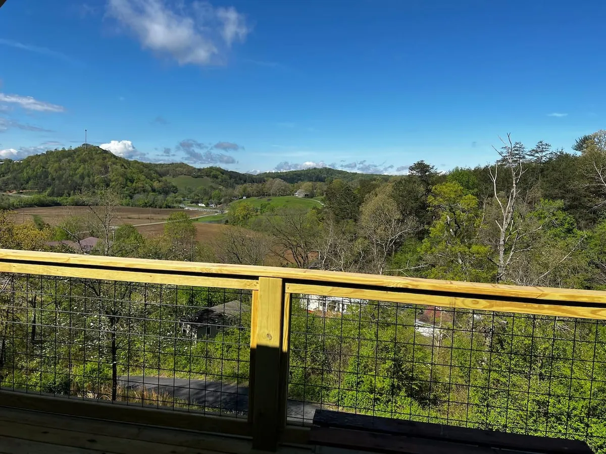 A sweeping view of a mountainous landscape with open fields under a vast blue sky, seen from a wooden deck railing. The scene suggests changing seasons with the potential for colorful autumn foliage, wildflower blooms, and lush greenery. The peaceful setting invites morning coffee, sunset watching, and wildlife observation.