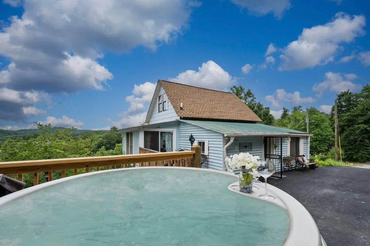 The image showcases a charming log cabin nestled in lush woodland, with a spacious deck overlooking sweeping Smoky Mountain vistas. A bubbling hot tub sits front and center, beside cozy outdoor seating and a dining area, all perfectly positioned to take in sunrise or sunset hues over the mountains.