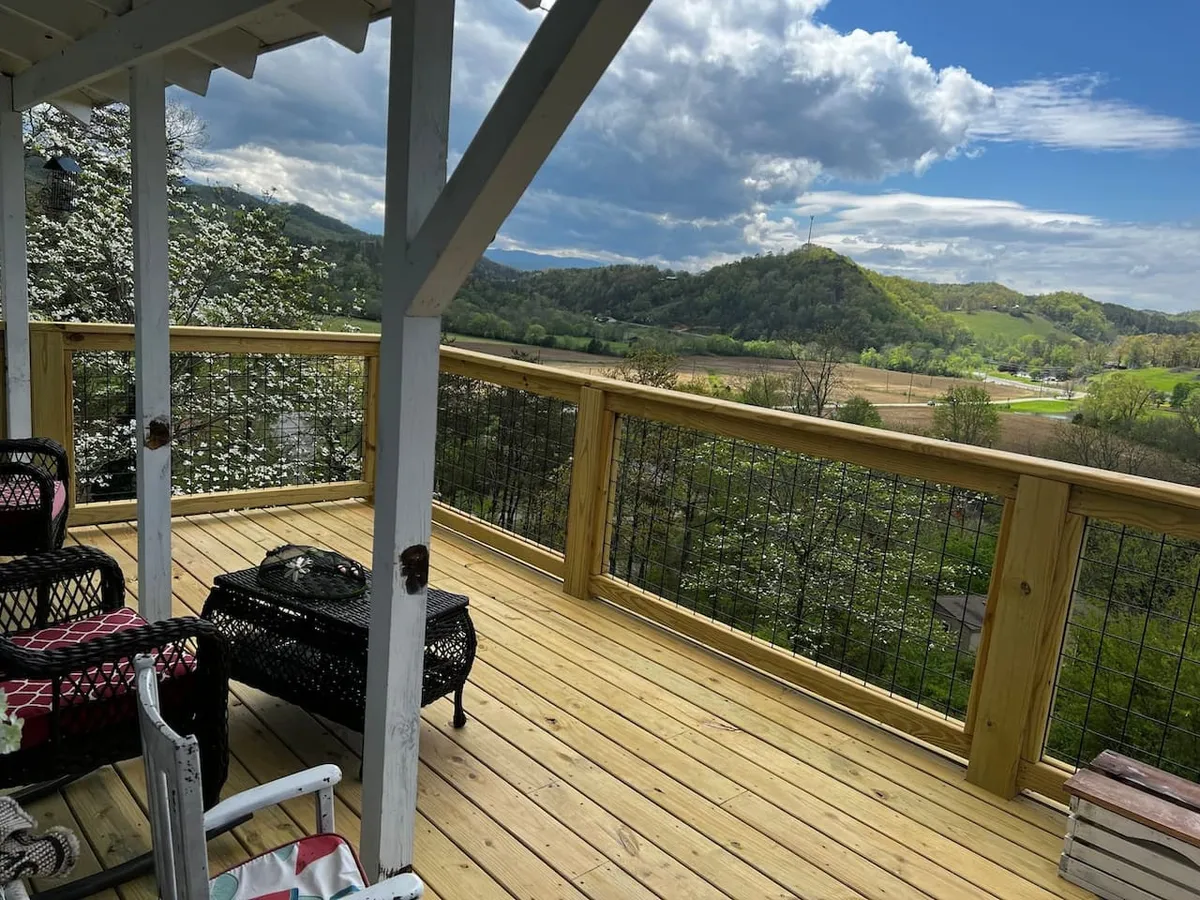 A spacious wooden deck furnished with comfortable outdoor furniture overlooks a stunning mountainous landscape. The deck invites relaxation, with ample space for rocking chairs and room to take in the sunrise or sunset, set against a tranquil natural backdrop.